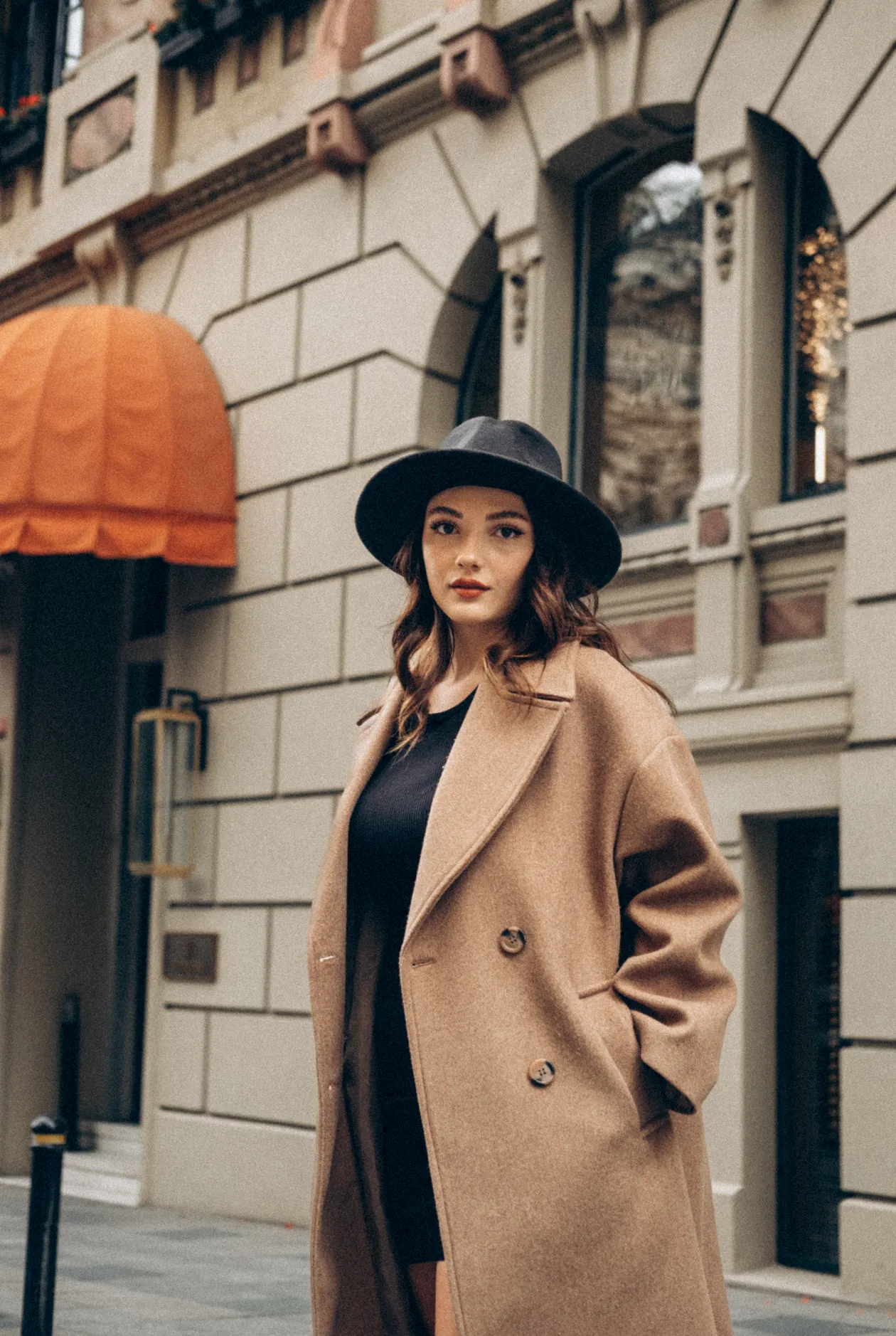 Woman in a camel coat over a black top with a fedora hat on an urban street.