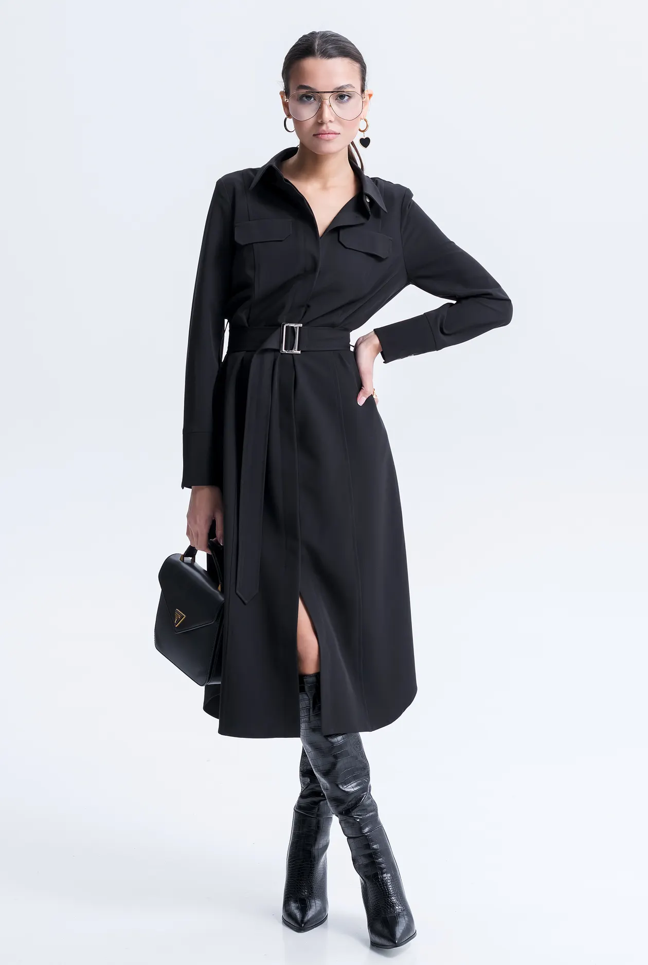 Woman in a black midi shirt dress with a slim belt, boots, and a handbag in a white studio.