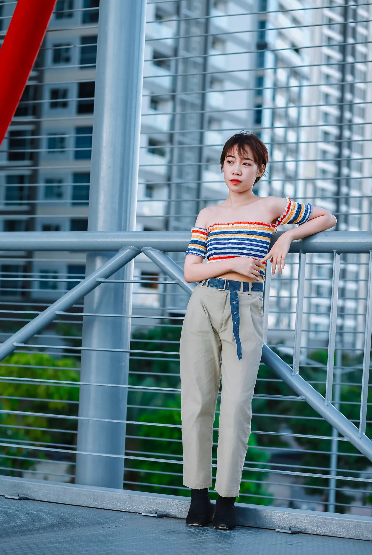 Woman in a striped off-shoulder crop top and beige high waist chinos with a blue belt with a long end hanging, on an urban bridge.