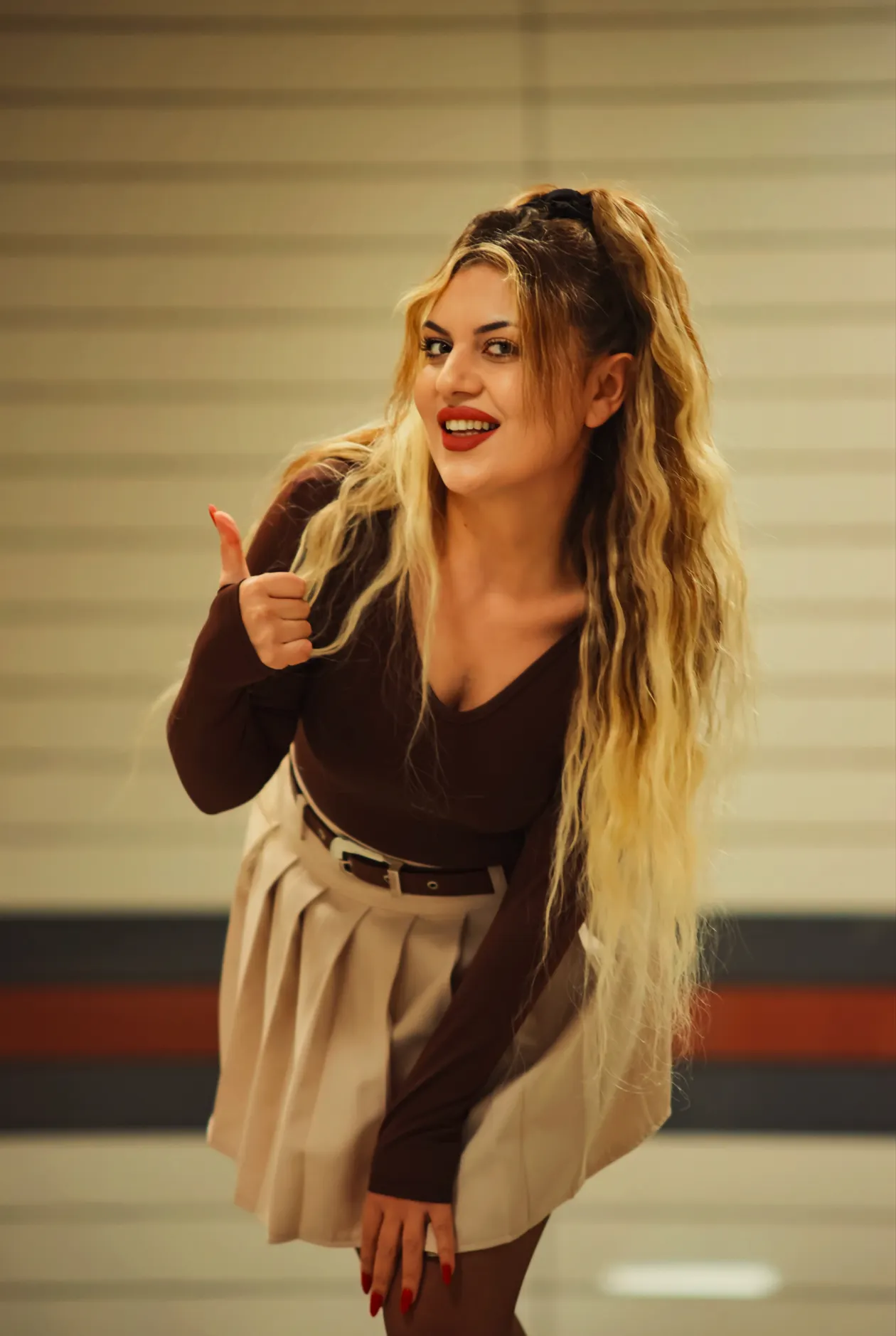 Woman in a fitted brown top and a beige pleated skirt with a dark belt, giving a thumbs-up indoors.