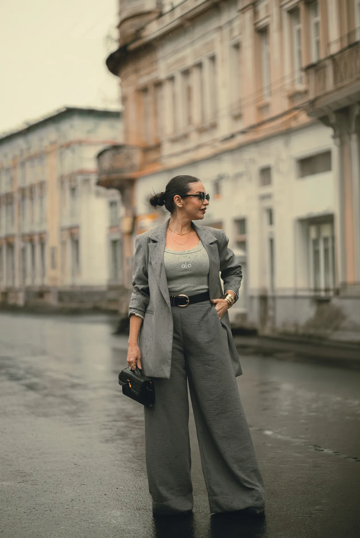 Woman in a grey oversized blazer with a black belt with a gold buckle and wide leg grey trousers on a wet city street.