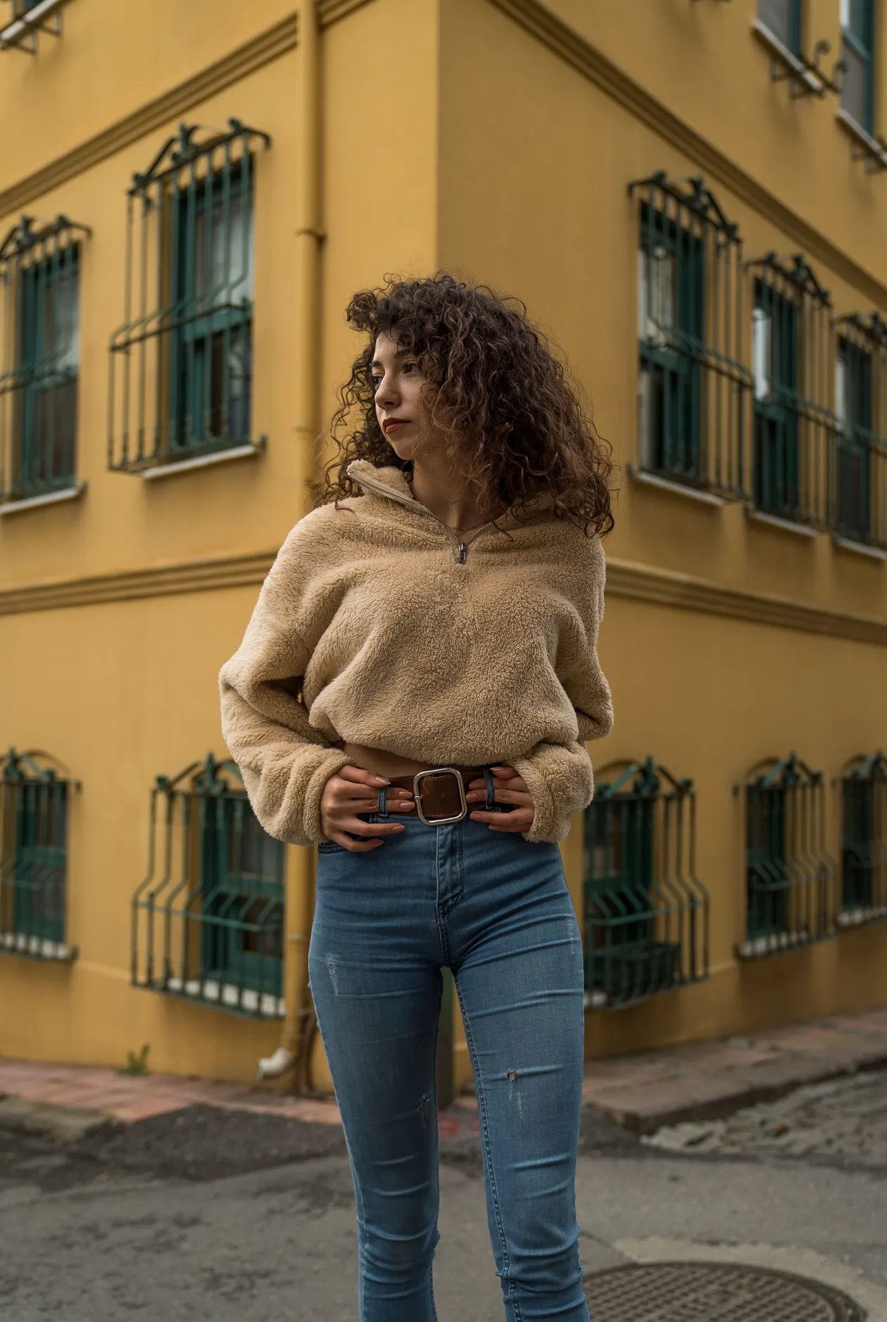 Woman in a tan fleece sweater, blue jeans, and a wide brown leather belt standing on a street by a yellow building.