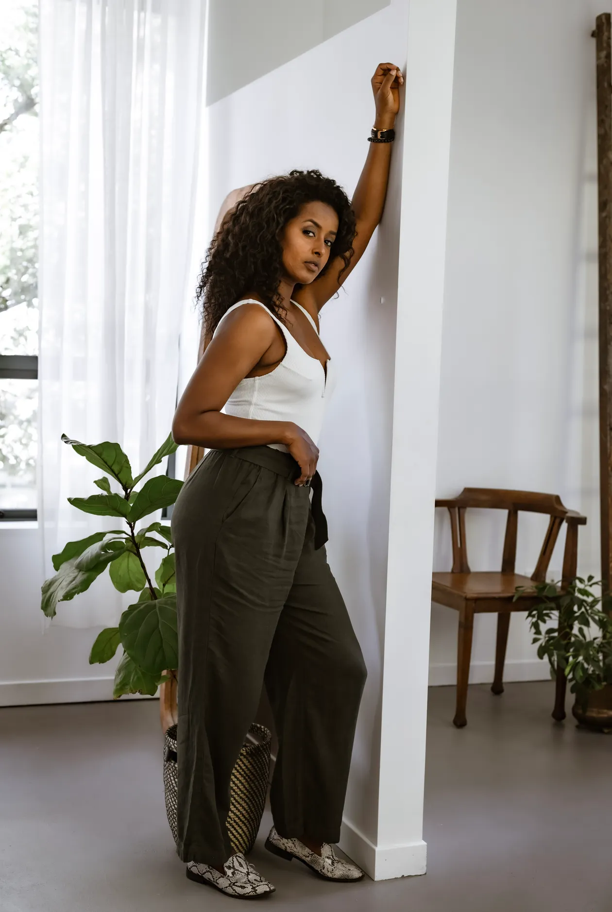 Woman in a white tank top and olive paperbag waist pants with a tie belt in a modern interior.