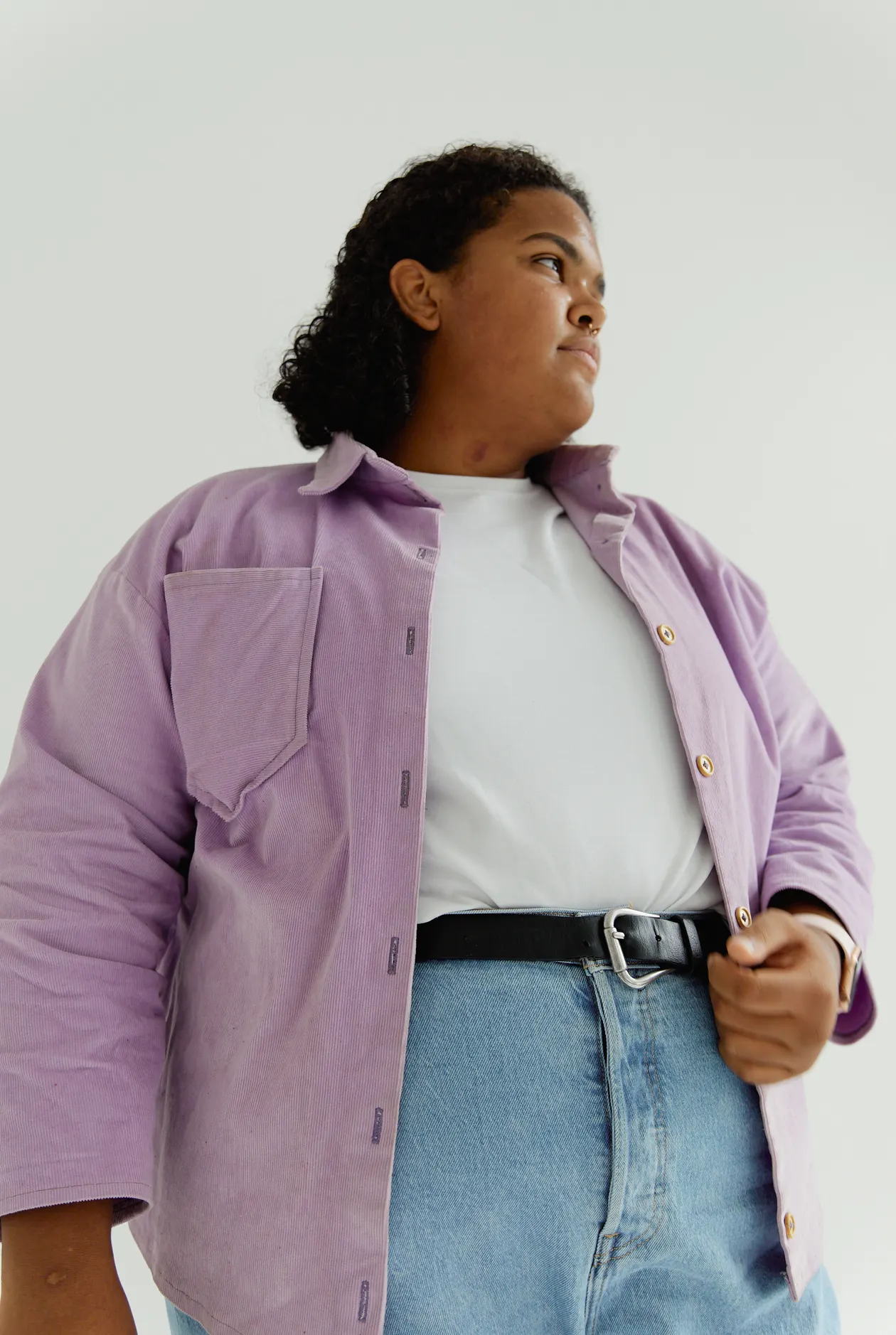 Woman in a lilac corduroy overshirt over a white top, blue jeans, and a black belt.