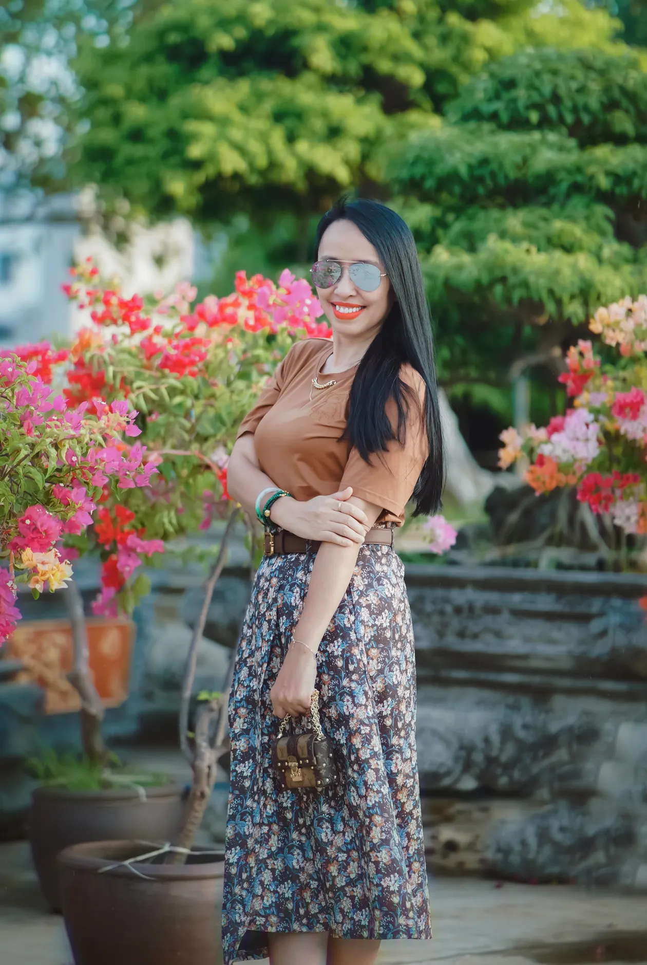 Woman in a brown top and a floral midi skirt with a brown belt partly hidden by her arm, in a garden.