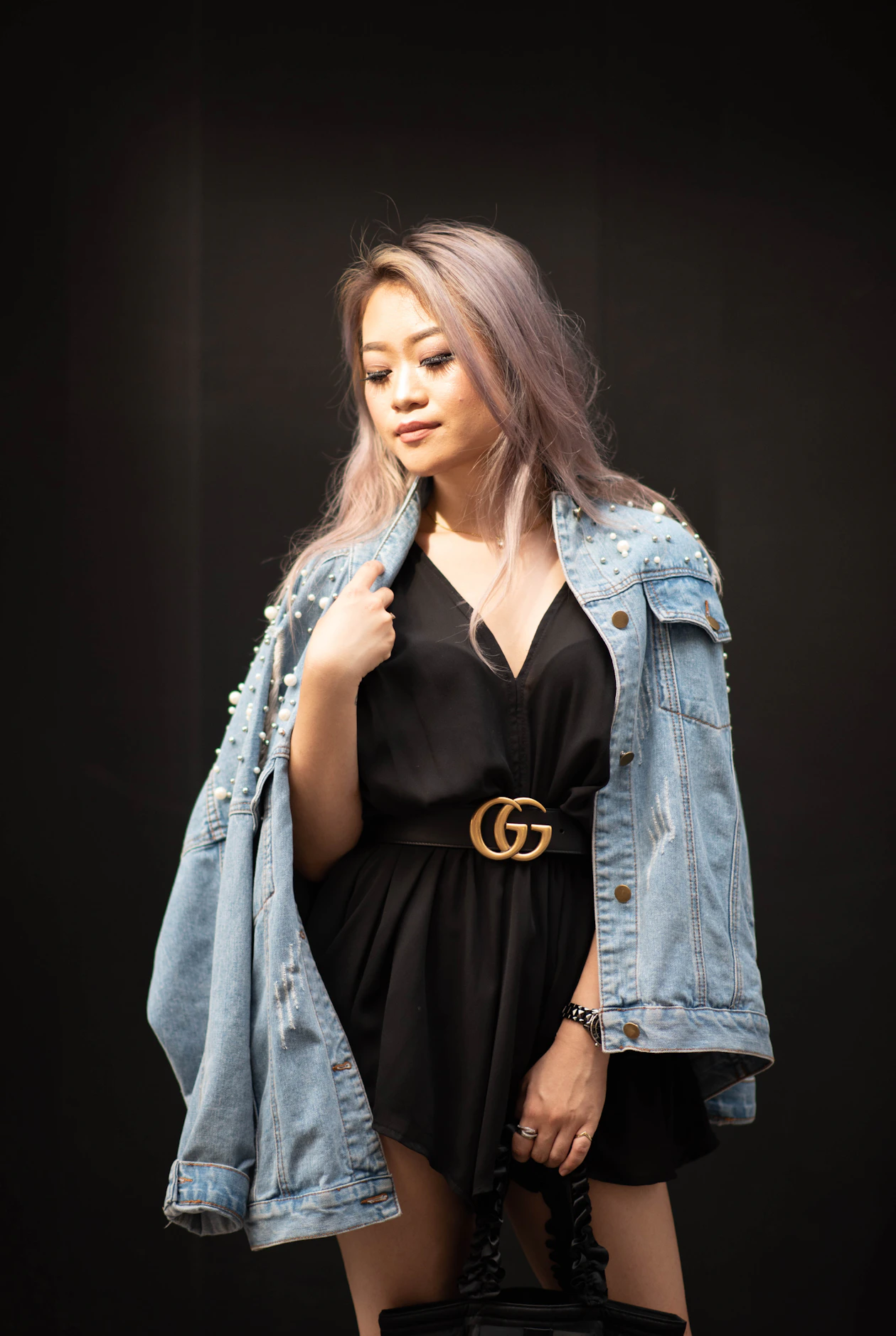 Woman in a black dress with a pearl-studded light denim jacket open and a black belt with a gold double-G buckle.