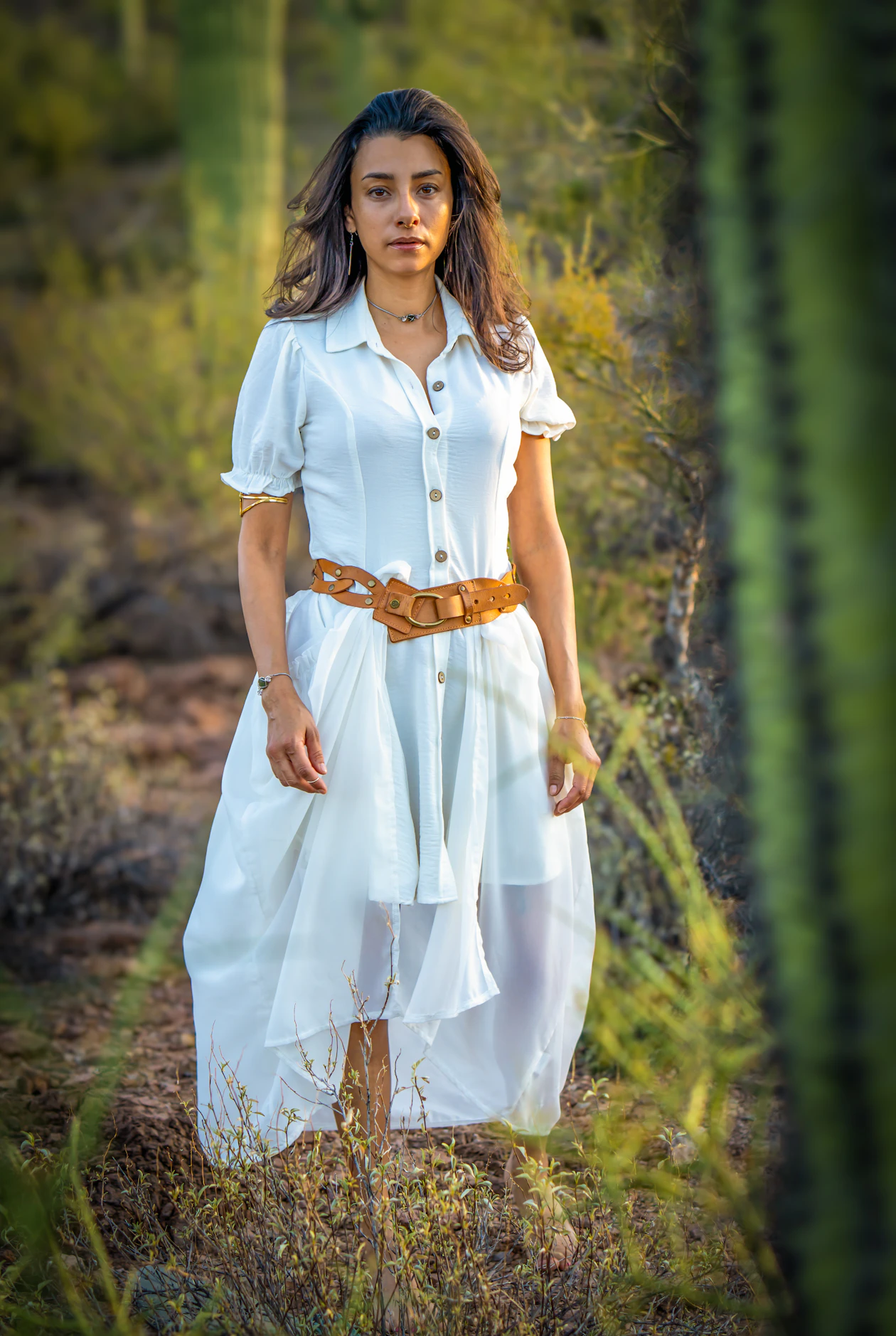 Woman in a white shirt dress with a wide tan belt with woven-style cutout detailing, standing in a cacti field.